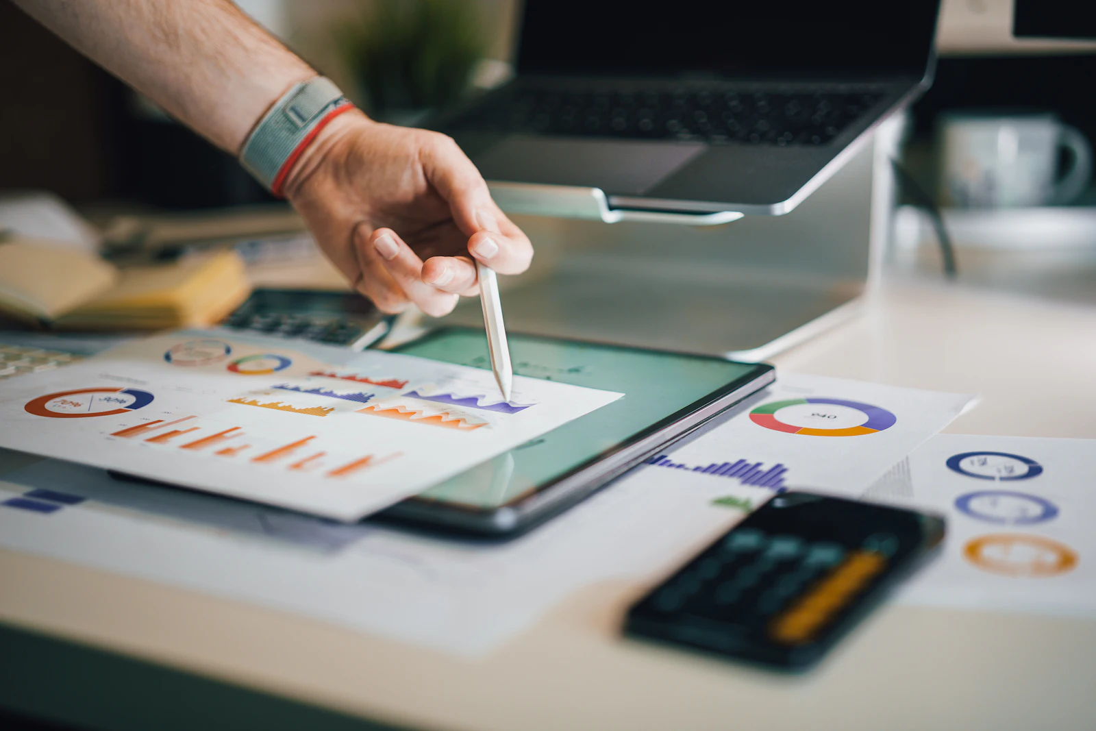 Hand with a stylus marking up printed charts beside a tablet